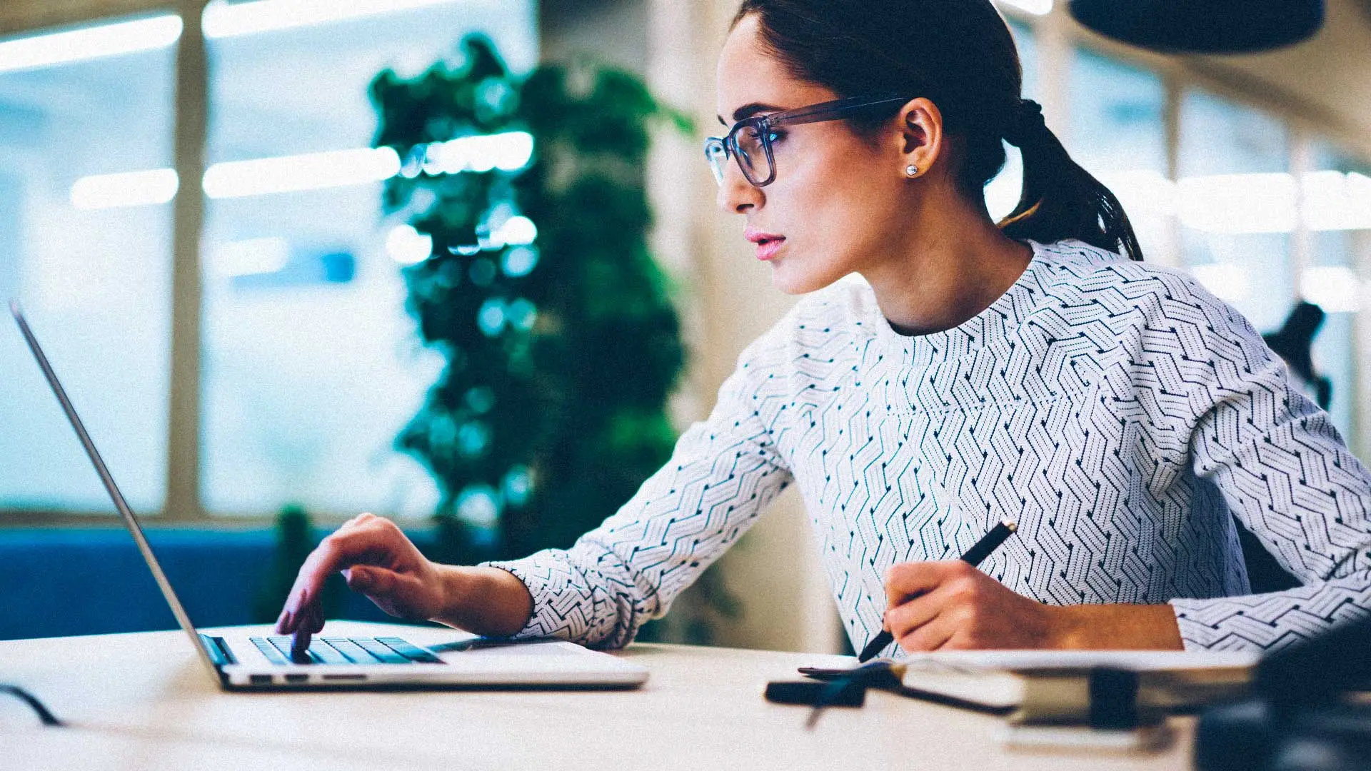 A woman sits at a desk looking at a laptop while holding a pen near a notebook.