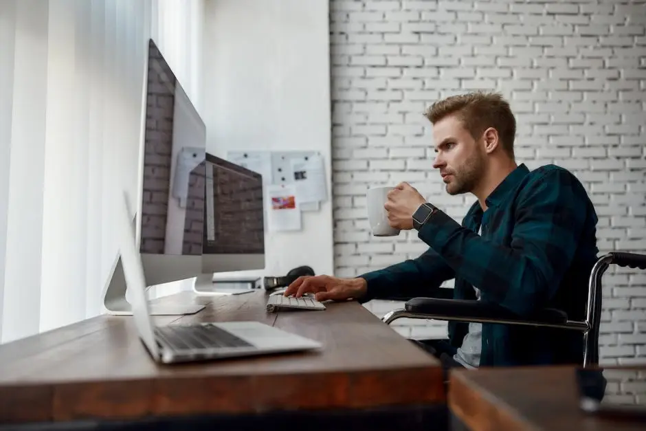 Matin chargé.
Vue latérale d’un jeune développeur Web dans un fauteuil roulant codant sur plusieurs écrans d’ordinateur et buvant du café alors qu’il était assis sur son lieu de travail dans le bureau moderne 