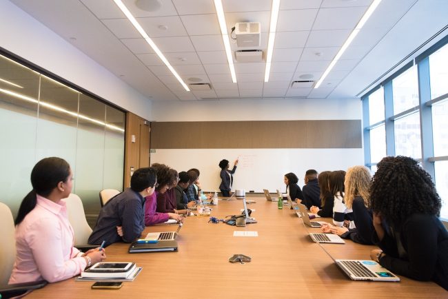 A group of professional adults sit around a table in a conference room setting. They are looking in the direction of the front of the room where a Black woman is presenting and writing on a whiteboard.