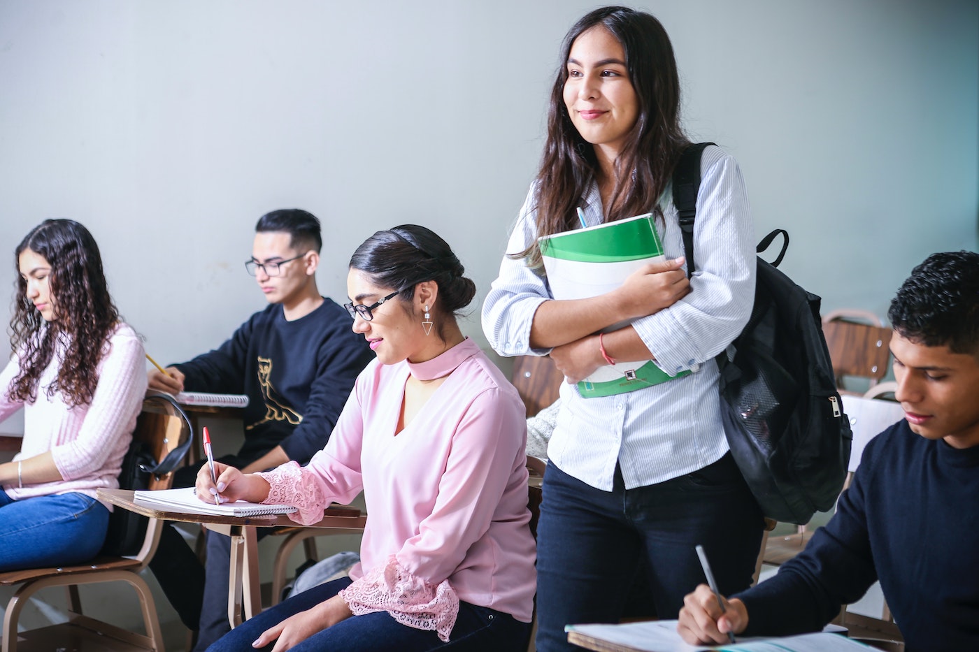 Five diverse students are in a classroom setting. Four of the students are happily sitting at their desks, writing in notebooks. One student, a young female with long dark hair and olive skin tone is holding her notebook in her arms and smiling the direction of the front of the classroom.
