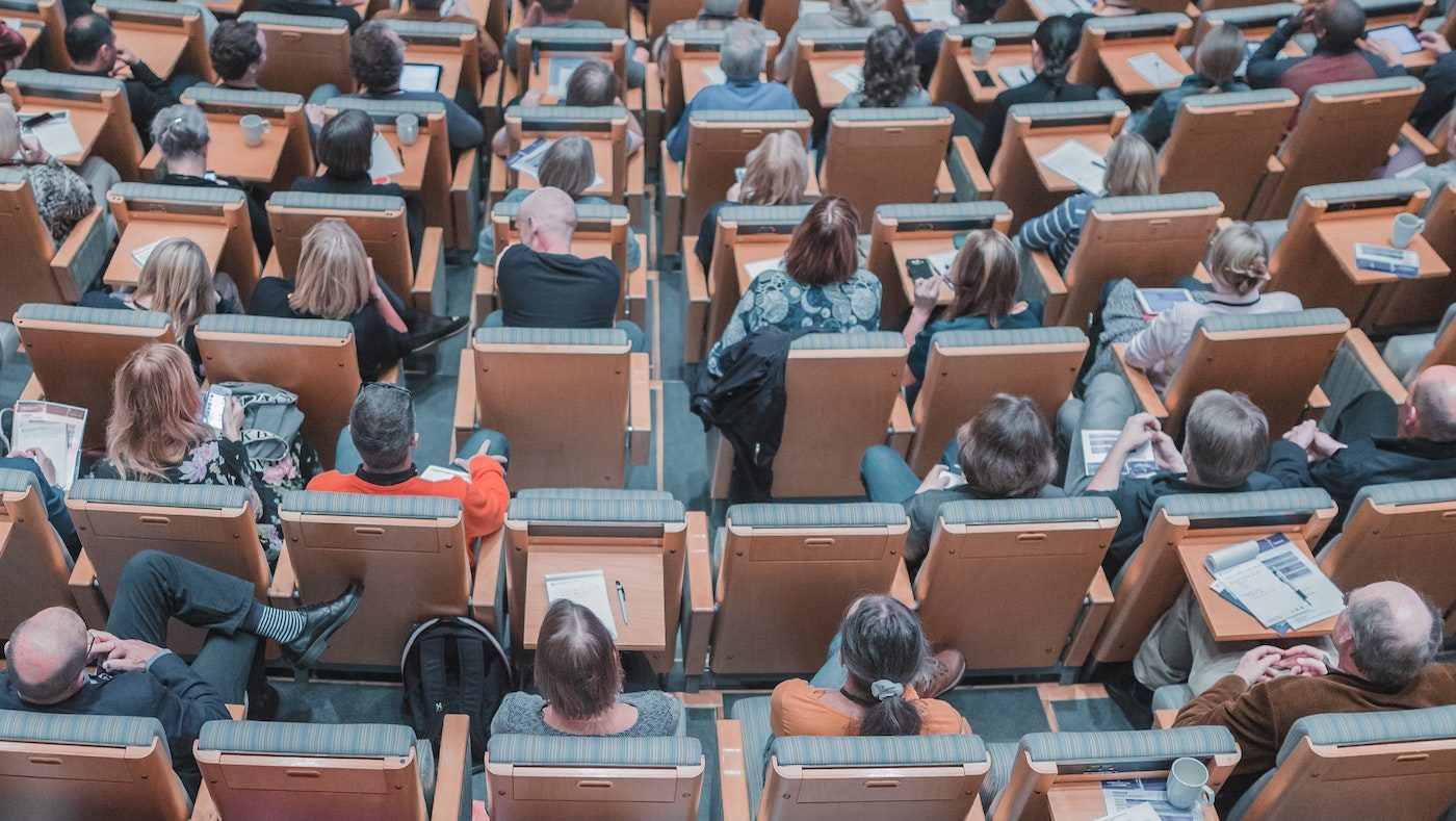 Aerial view of a lecture hall, in the wooden-backed seats are the students who range in age, gender, and race. They are all looking towards the front of the room, some are writing down notes.